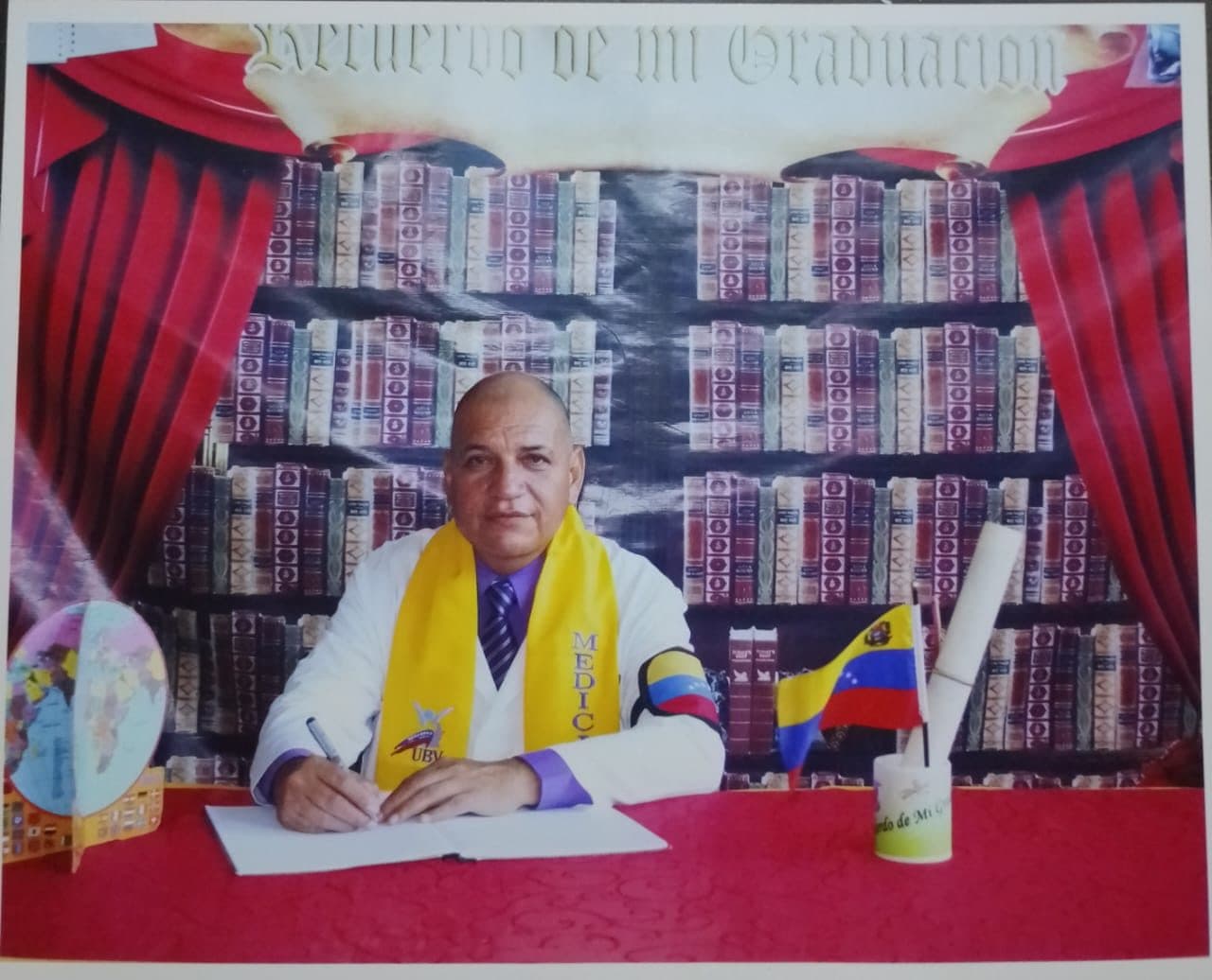 Fotografía de graduación del Dr. Iván López con estola amarilla, firmando sobre un escritorio con la bandera de Venezuela
